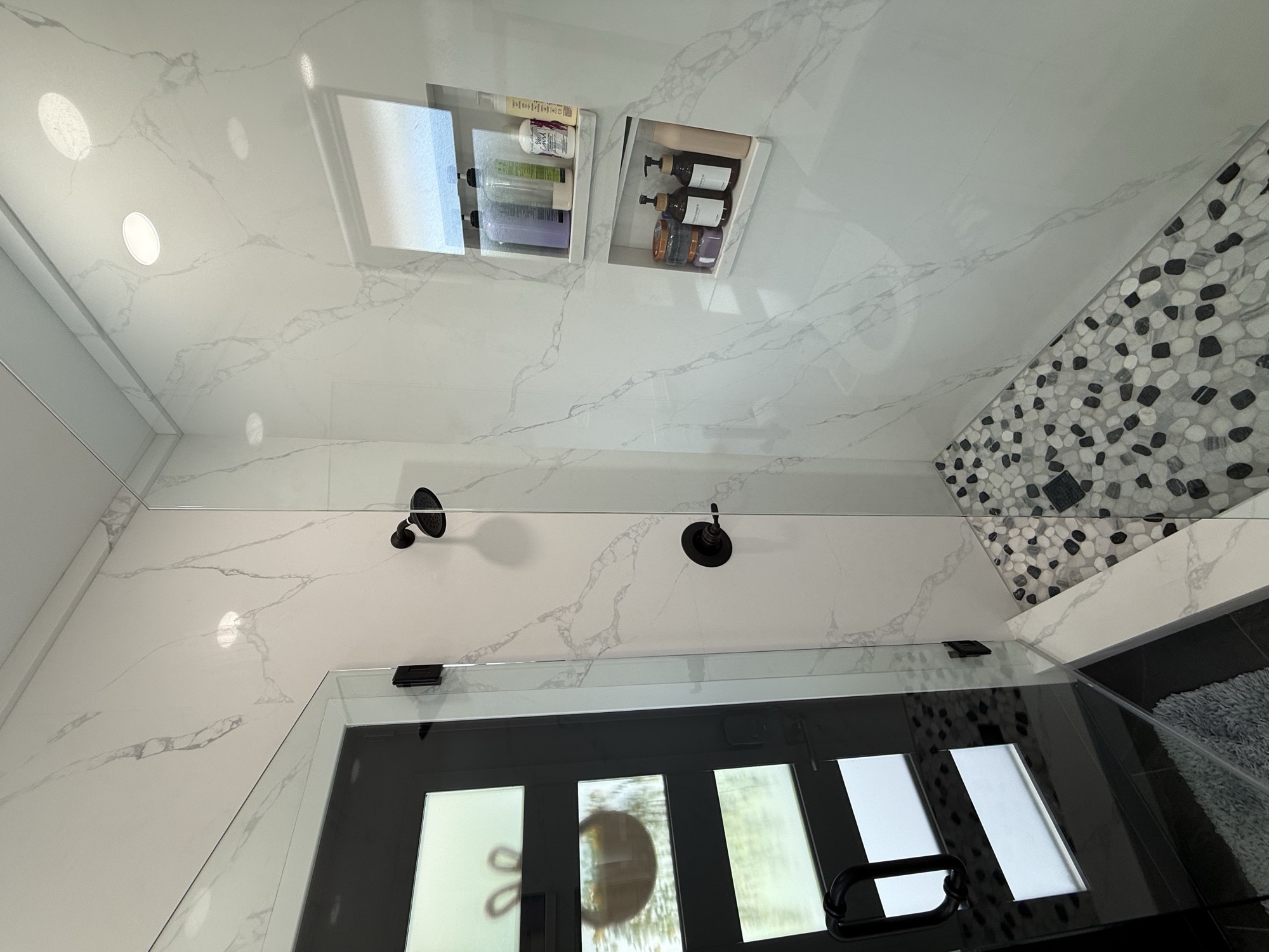 Another view of the same bathroom: glass shower enclosure, recessed niches, black fixtures, and pebble-pattern floor