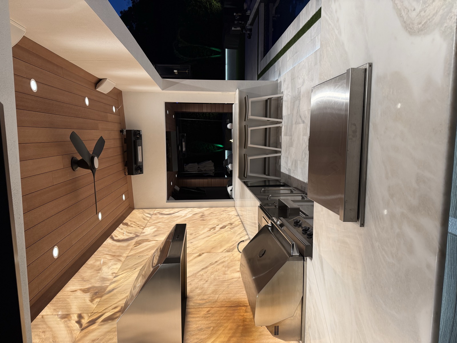 Covered outdoor kitchen with a long countertop, grill, bar seating, and warm stone wall
